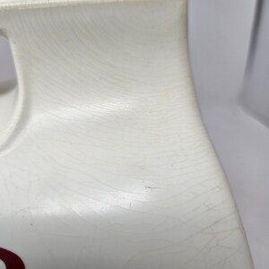 Puede incluir: Jarra de cer&aacute;mica blanca con una superficie texturizada y un dise&ntilde;o rojo. La jarra tiene un asa y un pico.