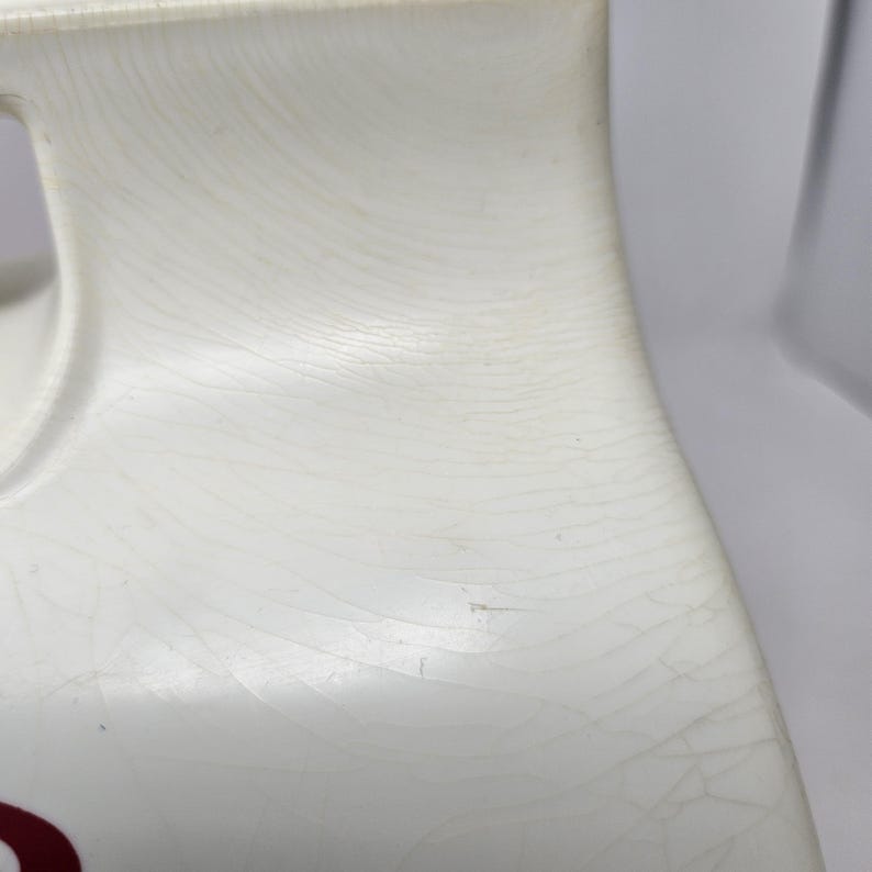 Puede incluir: Jarra de cer&aacute;mica blanca con una superficie texturizada y un dise&ntilde;o rojo. La jarra tiene un asa y un pico.