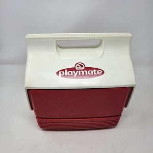 Puede incluir: Una nevera Igloo Playmate blanca y roja con una manija roja y una tapa roja. La nevera tiene el logotipo de Igloo y la palabra "Playmate" impresa en la parte delantera.
