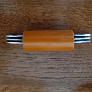 May include: A close-up of a metal tool with three blades attached to a cylindrical orange plastic handle.