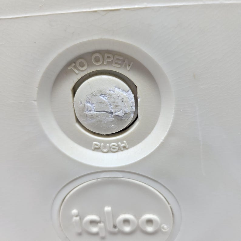 Puede incluir: Primer plano de una nevera blanca con un bot&oacute;n redondo blanco que dice "TO OPEN" y "PUSH". La nevera tiene una etiqueta blanca que dice "igloo."