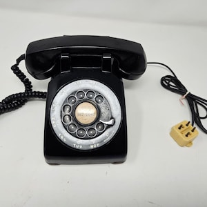 May include: A black rotary dial phone with a black coiled cord and a yellow modular jack. The phone has a silver dial with the numbers 362-2042 printed on it.