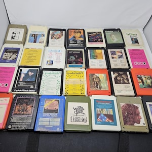 May include: A collection of vintage 8-track tapes, featuring various album covers with images of musicians and album art. The tapes are in different colors, including orange, yellow, and black, and are arranged on a white surface.