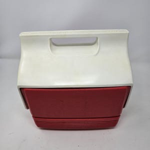 Puede incluir: Una nevera vintage roja y blanca con un asa en la parte superior. La nevera est&aacute; cerrada y tiene un fondo texturizado.