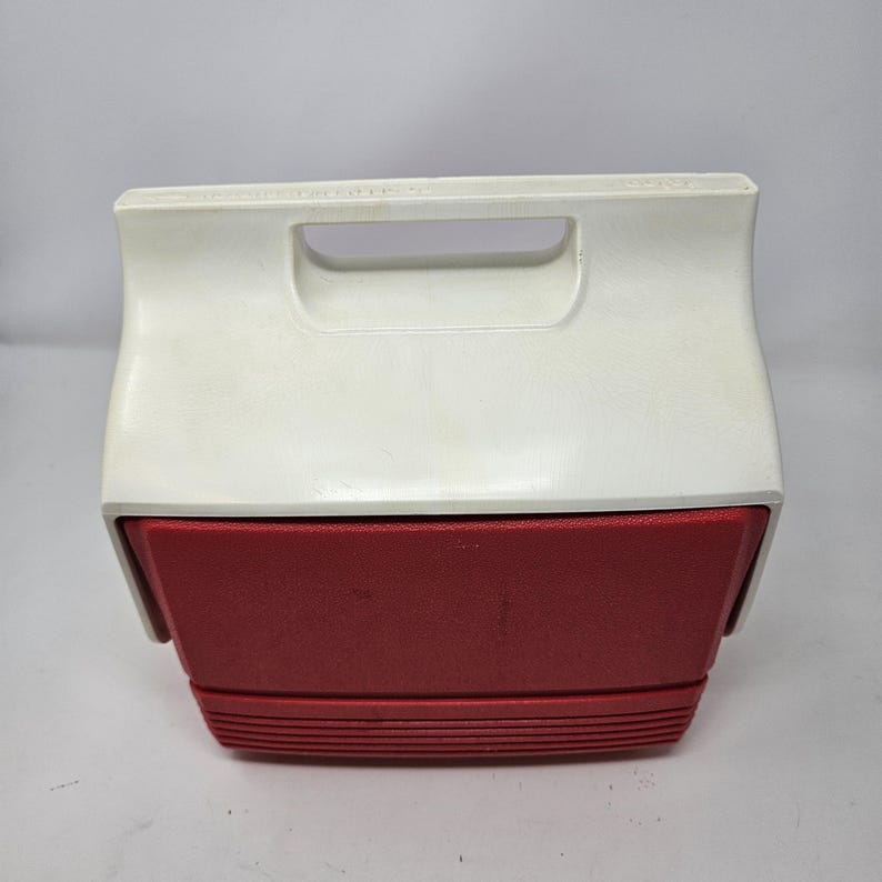 Puede incluir: Una nevera vintage roja y blanca con un asa en la parte superior. La nevera est&aacute; cerrada y tiene un fondo texturizado.