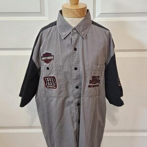 May include: A gray short-sleeve button-down shirt with black sleeves and collar accents. The shirt features embroidered Harley-Davidson logos and text on the chest pockets and sleeves, including the Harley-Davidson Motor Cycles shield logo.