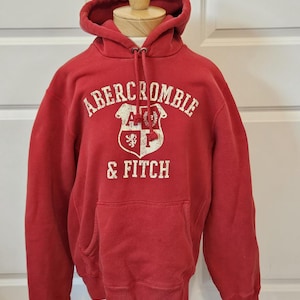 May include: A red hooded sweatshirt featuring a white distressed graphic print on the chest. The text reads ABERCROMBIE & FITCH above and below a shield emblem containing the letters A and F, a lion, and a laurel wreath. It has a front kangaroo pocket.