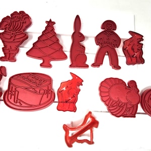 May include: A collection of red plastic cookie cutters in various shapes, including Santa Claus, a Christmas tree, a rabbit, a person, a pig, a cake, a witch, a turkey, a jack-o'-lantern, and a megaphone. These cutters are ideal for making festive cookies.