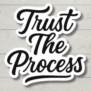 May include: A white sticker with the words "Trust The Process" in black cursive font. The sticker has a black border and is set against a light wood grain background.
