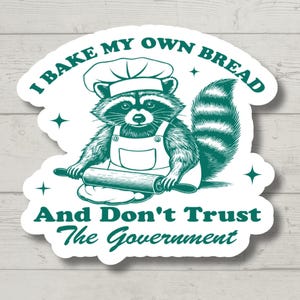 May include: A green and white sticker with a cartoon raccoon wearing a chef's hat and holding a rolling pin. The text reads "I bake my own bread and don't trust the government."
