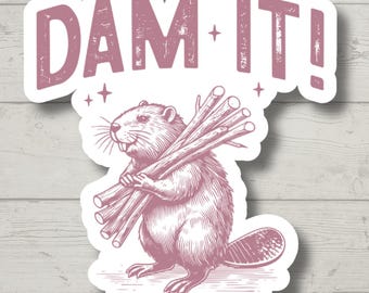 Op de afbeelding: Een sticker met een roze illustratie van een bever die boomstammen draagt, met de tekst "DAM IT!". Het ontwerp heeft een verweerde, vintage uitstraling, met een witte rand. Houten achtergrond.