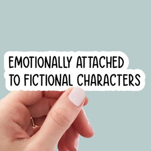 May include: A white die-cut sticker with a black border and the black text "EMOTIONALLY ATTACHED TO FICTIONAL CHARACTERS" written in a rounded sans-serif font. The sticker features a clean, minimalist design with a matte finish and a white outline.
