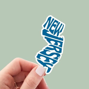May include: A blue and white sticker in the shape of the state of New Jersey. The word "NEW JERSEY" is printed in a stylized font within the state's outline. The sticker is held by a hand against a light green background.