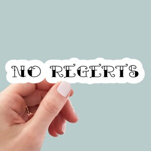 May include: A white sticker with black text that reads "NO REGRETS" in a decorative font. A hand is holding the sticker.