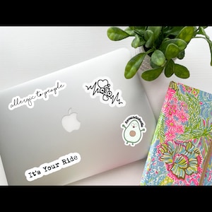 May include: A silver laptop with four stickers on it. The stickers say "allergic to people", "It's Your Ride", "Avocadoholic", and a heart with a heartbeat line.