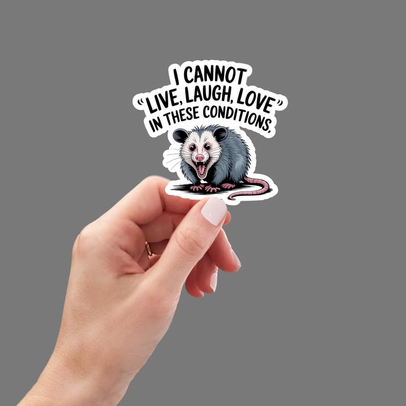 May include: A sticker featuring a cartoon opossum with a wide open mouth and the text "I CANNOT 'LIVE, LAUGH, LOVE' IN THESE CONDITIONS." The opossum is gray, white, and pink, with a black outline. The sticker is held by a hand.