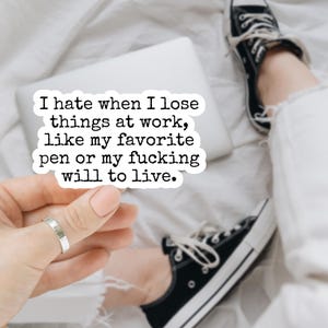 May include: A white sticker with black text that reads, "I hate when I lose things at work, like my favorite pen or my fucking will to live." The sticker is held by a hand with a silver ring. A laptop, black sneakers, and white pants are in the background.