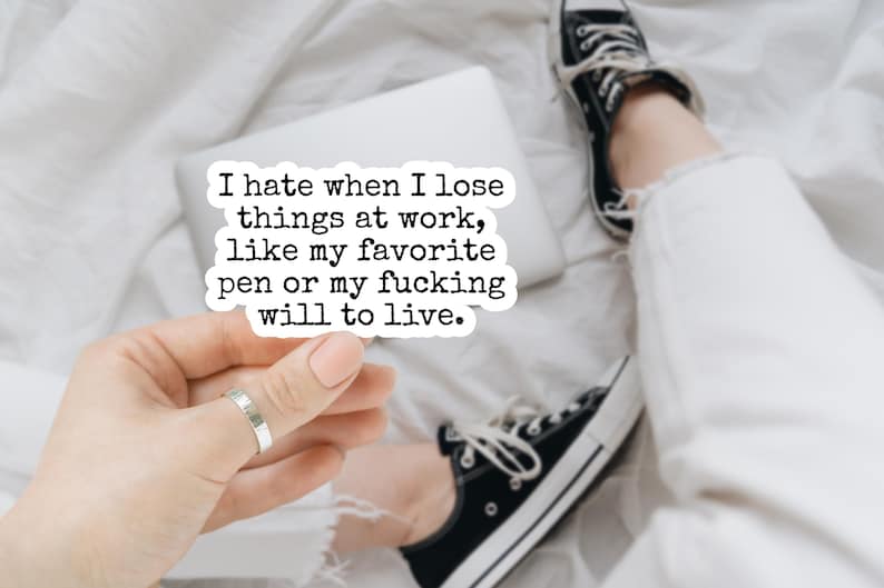 May include: A white sticker with black text that reads, "I hate when I lose things at work, like my favorite pen or my fucking will to live." The sticker is held by a hand with a silver ring. A laptop, black sneakers, and white pants are in the background.