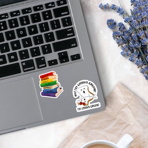May include: Two vinyl stickers on a laptop. One sticker shows a stack of colorful books with the text YOU BAN A BOOK I ADD IT TO MY SUMMER READS LIST. The other sticker features two slices of bread with the text OH! I'M GONNA CRUMB! I'M GONNA CRUMB!