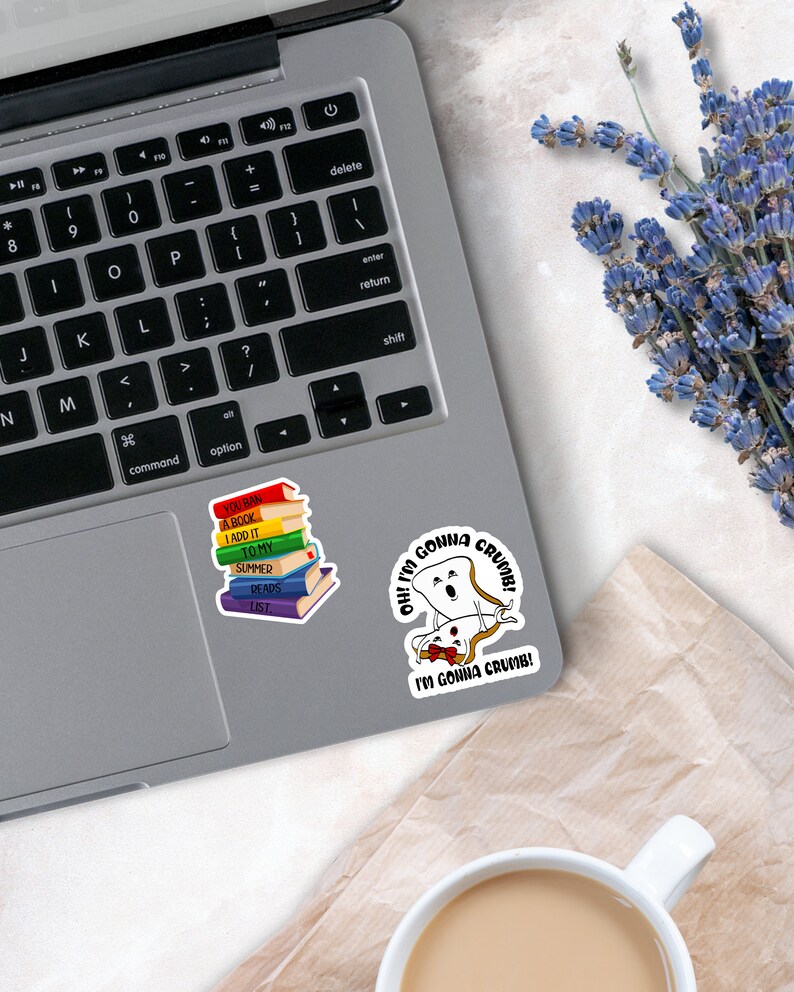 May include: Two vinyl stickers on a laptop. One sticker shows a stack of colorful books with the text YOU BAN A BOOK I ADD IT TO MY SUMMER READS LIST. The other sticker features two slices of bread with the text OH! I'M GONNA CRUMB! I'M GONNA CRUMB!