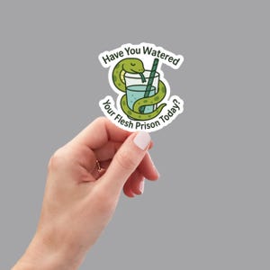 May include: A sticker featuring a cartoon snake coiled around a glass of water with a straw. The text reads "Have You Watered Your Flesh Prison Today?" in a circular design. The sticker is held by a hand against a gray background.
