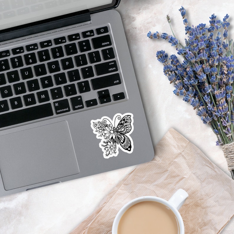 May include: Black and white sticker of a butterfly with floral details on a silver laptop.