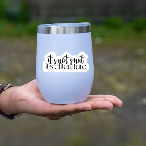 May include: A light lavender stainless steel tumbler with a silver rim and a clear lid. The front features a white sticker with black cursive text that reads: it's not smut it's cliterature. The tumbler has a smooth, rounded, stemless design.