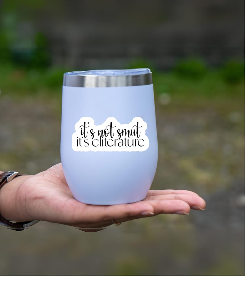 May include: A light lavender stainless steel tumbler with a silver rim and a clear lid. The front features a white sticker with black cursive text that reads: it's not smut it's cliterature. The tumbler has a smooth, rounded, stemless design.