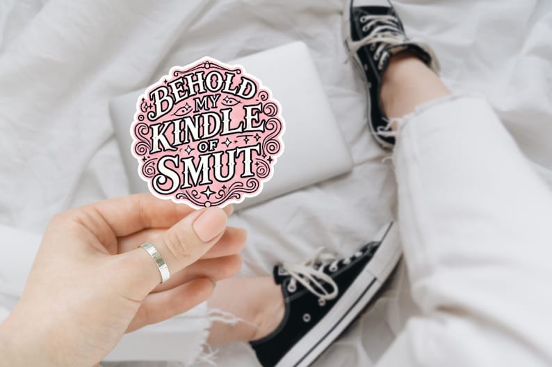 May include: A circular sticker with a light pink background and a decorative black border. The text reads BEHOLD MY KINDLE OF SMUT in bold, stylized black lettering, surrounded by intricate swirling flourishes and small star accents.