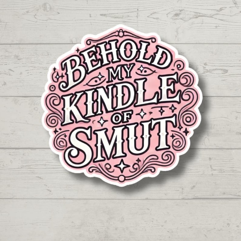 May include: A circular sticker with a light pink background and a decorative black border. The text reads BEHOLD MY KINDLE OF SMUT in bold, stylized black lettering. Small black star and eye icons are interspersed throughout the design.
