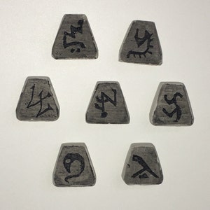 May include: Seven grey, trapezoid-shaped wooden runes with black symbols. The runes are arranged in a circular pattern. Each rune has a unique symbol, possibly used for divination or spiritual practices.