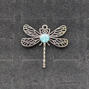 May include: Silver dragonfly charm with a blue and white patterned glass center. The dragonfly has intricate details and a long, beaded tail.
