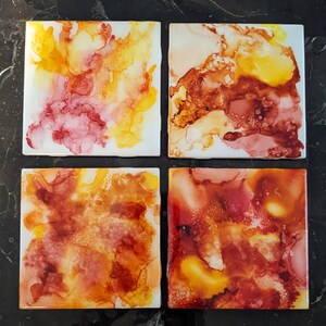 May include: Set of four square ceramic coasters featuring abstract watercolour designs in shades of yellow, orange, red, and brown. The coasters are ideal for protecting surfaces from heat and spills.