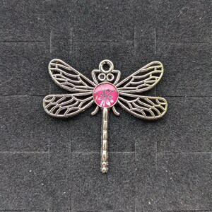 May include: Silver dragonfly charm with a pink glass cabochon in the center. The cabochon has a small dragonfly design inside.