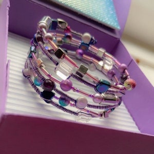 Könnte beinhalten: Ein mehrsträngiges Drahtarmband mit Perlen in Lila, Rosa und schillernden Farben. Das Armband wird in einer lila Geschenkbox präsentiert, die sein aufwendiges Design und seine Farbpalette hervorhebt. Maße in Zentimetern.