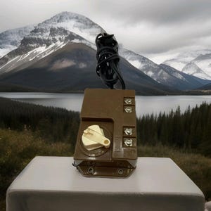 May include: A brown electrical transformer with a cream-colored dial and a black power cord. The transformer has several connection points and is sitting on a white surface. The background features a scenic view of mountains and a lake.