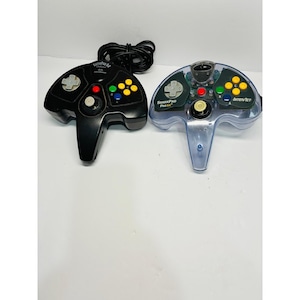 May include: Two video game controllers for the N64 console. One is solid black with the text SuperPad 64, and the other is transparent blue with the text SharkPad Pro 64 and InterAct. Both feature a central analog stick, a directional pad, and four colored action buttons labeled A, B, and C.
