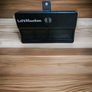 May include: A black LiftMaster garage door opener with three rectangular buttons and a metal loop. The LiftMaster logo and a circular design are visible on the front of the device. The opener is set against a wooden background.