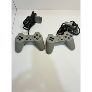 May include: Two original grey Sony PlayStation game controllers with wired connections and grey rectangular plugs. Each controller features a directional pad, four action buttons with colored symbols, start and select buttons, and the Sony PlayStation logo.