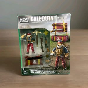 May include: A Mega Construx Call of Duty Armored Division Weapon Crate set. The box features two soldier figures, weapons, and crates. The figures are wearing red and tan uniforms. The box has the text "Call of Duty" and "Armored Division Weapon Crate".