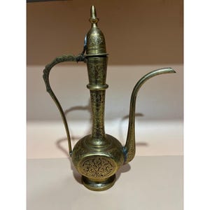 Puede incluir: Una jarra de agua antigua de latón con intrincados grabados florales y geométricos. La jarra presenta un pico largo y curvo, un asa decorativa y una tapa con bisagras. El diseño general es ornamentado y detallado, mostrando una estética vintage.