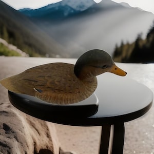 Könnte beinhalten: Eine hölzerne Entenattrappe mit braunen und weißen Federn, die auf einem schwarzen Tisch mit einem verschwommenen Hintergrund aus Bergen und Bäumen steht.