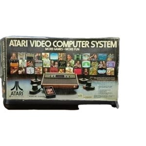 Puede incluir: Un juego de caja del sistema Atari Video Computer. La caja presenta el logotipo de Atari y el texto "MÁS JUEGOS - MÁS DIVERSIÓN". El juego incluye una consola, mandos y cartuchos de juego. La caja es marrón, negra y blanca.