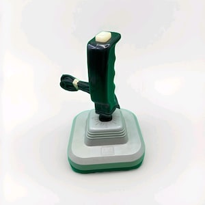 以下が含まれることがあります： 黒いハンドルと白いボタンが付いた、ヴィンテージのジョイスティックコントローラー。ベースはグレーとグリーンで、グリーンの縁取りがあります。黒いコードがジョイスティックに接続されています。