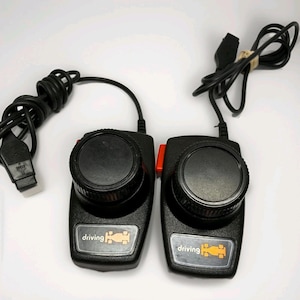 May include: Two black video game driving controllers with circular knobs and red side buttons. Each controller features a rectangular label at the base with the text driving and a yellow race car icon, connected by black cords with connectors.