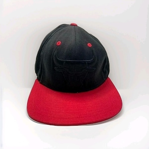 Puede incluir: Una gorra de béisbol negra con visera plana roja y un botón rojo en la parte superior. La parte frontal presenta un logotipo de toro negro bordado. La gorra tiene seis paneles con pequeños ojales de ventilación rojos en la corona.