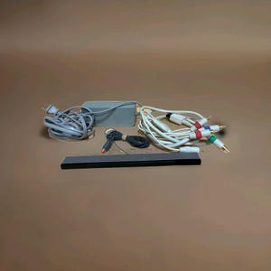 May include: A collection of electronic components, including a power adapter, cables with red, white, blue, and green connectors, and a black sensor bar. The items are arranged on a brown surface.