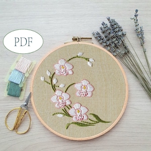 Puede incluir: Arte de bordado en aro con un delicado diseño floral de orquídeas blancas con acentos rosados sobre un fondo de lino beige.
