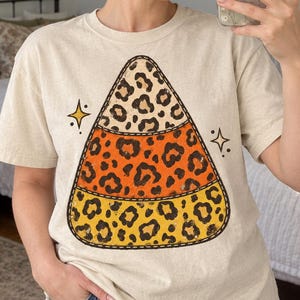 May include: A cream-coloured short-sleeve t-shirt featuring a large graphic of a candy corn shape divided into three horizontal sections, each filled with a black and brown leopard print pattern, accented by two small black star icons on the sides.