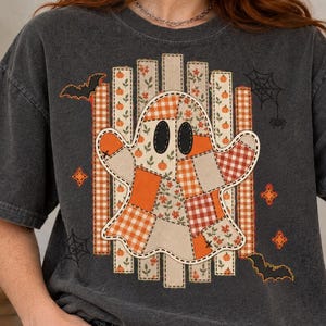 May include: A charcoal gray short-sleeve t-shirt featuring a central graphic of a ghost made of patchwork fabric patterns in orange, white, and brown. The design includes vertical stripes, small pumpkins, spiderwebs, and flying bats.